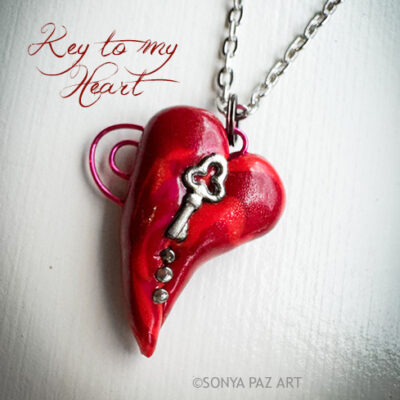 Red heart-shaped pendant with a silver key charm on a chain, against a light background, with the cursive words “Key to my Heart.”