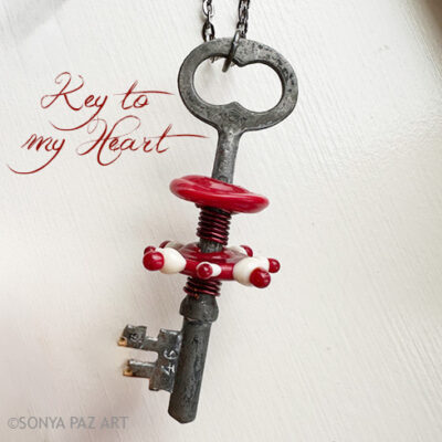 Old metal key with red beads and a red heart ring, hanging from a chain; the phrase "Key to my Heart" appears beside it.