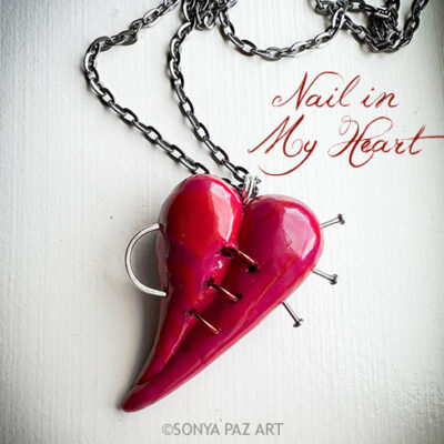 Pink heart-shaped pendant pierced with nails, hanging on a silver chain; caption reads 'Nail in My Heart'.