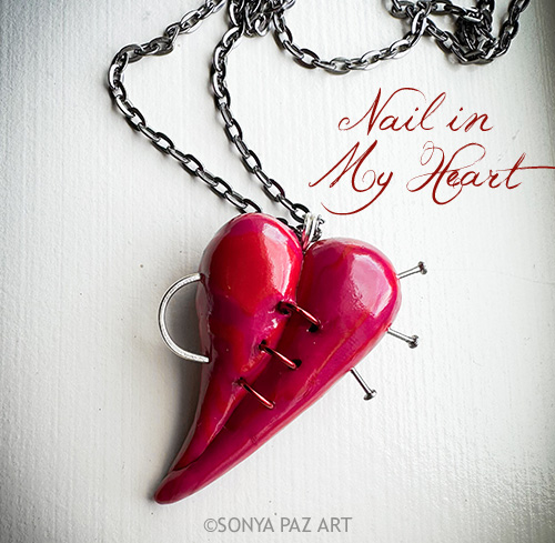 Pink heart-shaped pendant pierced with nails, hanging on a silver chain; caption reads 'Nail in My Heart'.