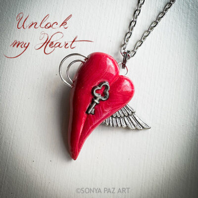 Red heart-shaped pendant with a key and wing charm on a chain, beside the handwritten phrase “Unlock my Heart.”
